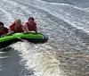 Three people are wearing life jackets excitedly riding a towable water tube behind a boat with coastal buildings in the background