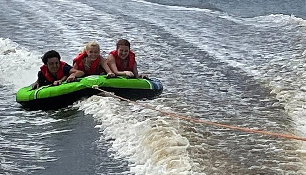 Three people wearing life jackets are riding a green inflatable tube being pulled over water creating a wake behind them