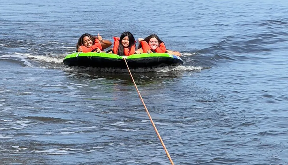 Three individuals wearing life jackets are riding on an inflatable tube towed by a boat on a body of water with one person giving a thumbs-up sign