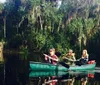 Three individuals are paddling in a green canoe through a serene waterway flanked by verdant trees draped with Spanish moss