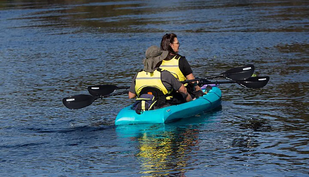 Two individuals are kayaking on calm water wearing life vests and using double-bladed paddles