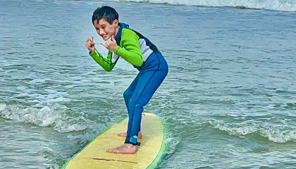 A young surfer wearing a green and blue wetsuit is balancing on a yellow surfboard riding a wave with a playful hand gesture