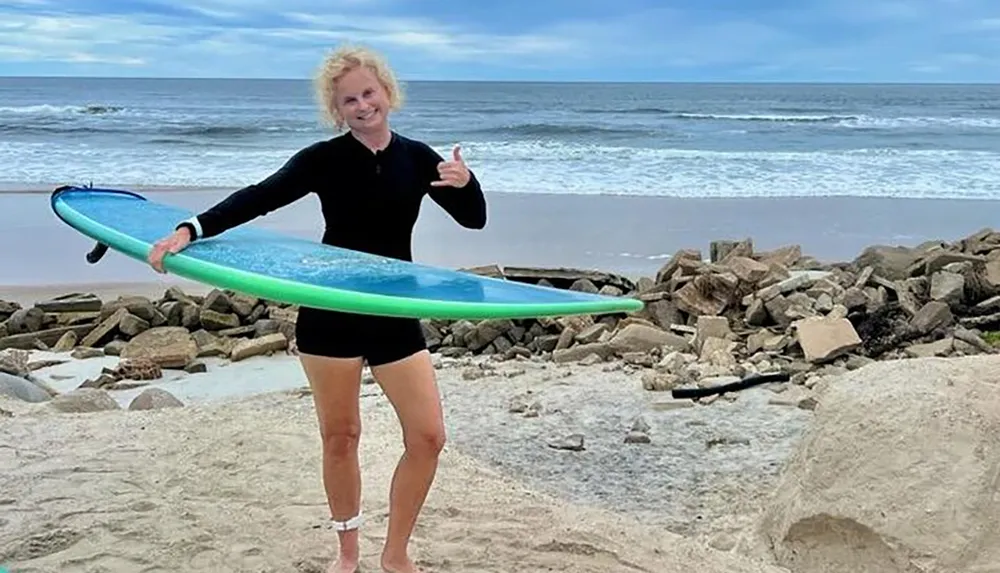 A person is smiling and giving a thumbs-up while holding a surfboard on a beach with waves in the background