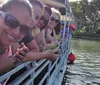A group of people wearing sunglasses and casual clothing are smiling and posing for a photo while seated on a boat with a railing backed by a scenic view of water and palm trees