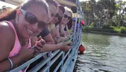 A group of people wearing sunglasses and casual clothing are smiling and posing for a photo while seated on a boat with a railing, backed by a scenic view of water and palm trees.