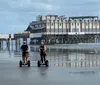 Two people are riding Segways on a wet beach with a large pier structure in the background