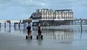 Two people are riding Segways on a wet beach with a large pier structure in the background.