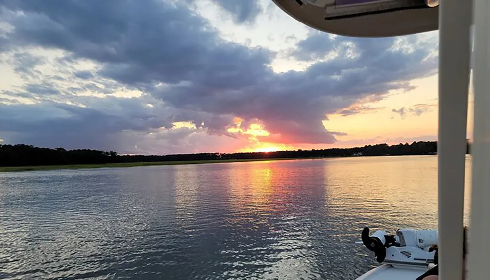 The image captures a serene sunset over a calm body of water viewed from a boat with visible parts of the vessel in the frame such as a section of railing and an outboard motor