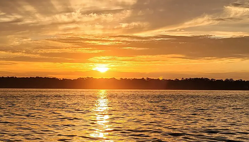 The image depicts a serene sunset with a radiant sun dipping towards the horizon casting a golden glow over the water and silhouetting the treeline