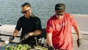 Two individuals are cooking food on a boat, one wearing a black chef's uniform and the other in a red tie-dye shirt with the phrase 