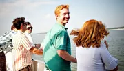 A group of people are enjoying a sunny day on a boat, with a man in a green polo shirt looking back at the camera with a smile.