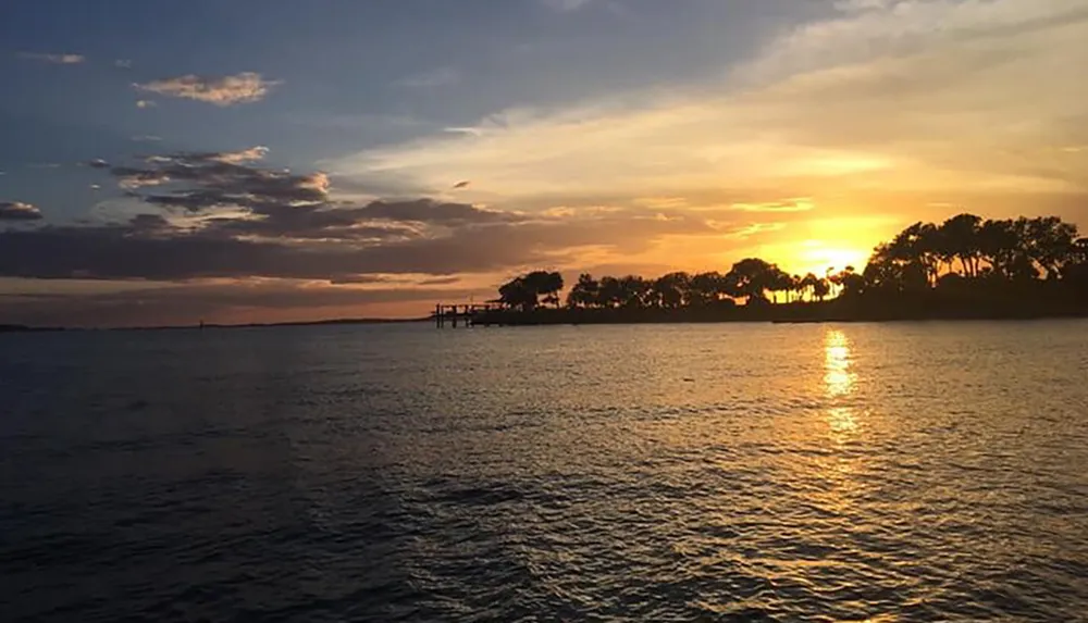 The image captures a serene sunset over a body of water with the suns golden reflection on the surface and silhouettes of trees on the horizon