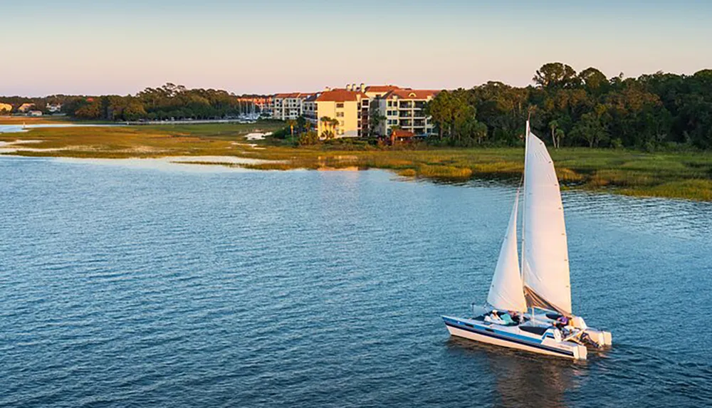 A sailboat is cruising near the shore with a backdrop of residential buildings and a lush treeline under a soft evening light