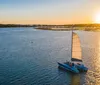 The image captures a sailboat gliding on calm waters with the sun setting in the background casting a golden reflection on the waters surface