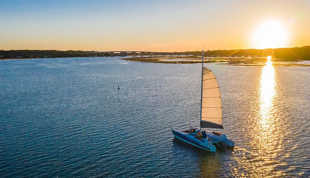 The image captures a sailboat gliding on calm waters with the sun setting in the background casting a golden reflection on the waters surface