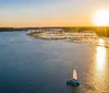 A sailboat glides across calm waters near a coastal settlement during a tranquil sunset