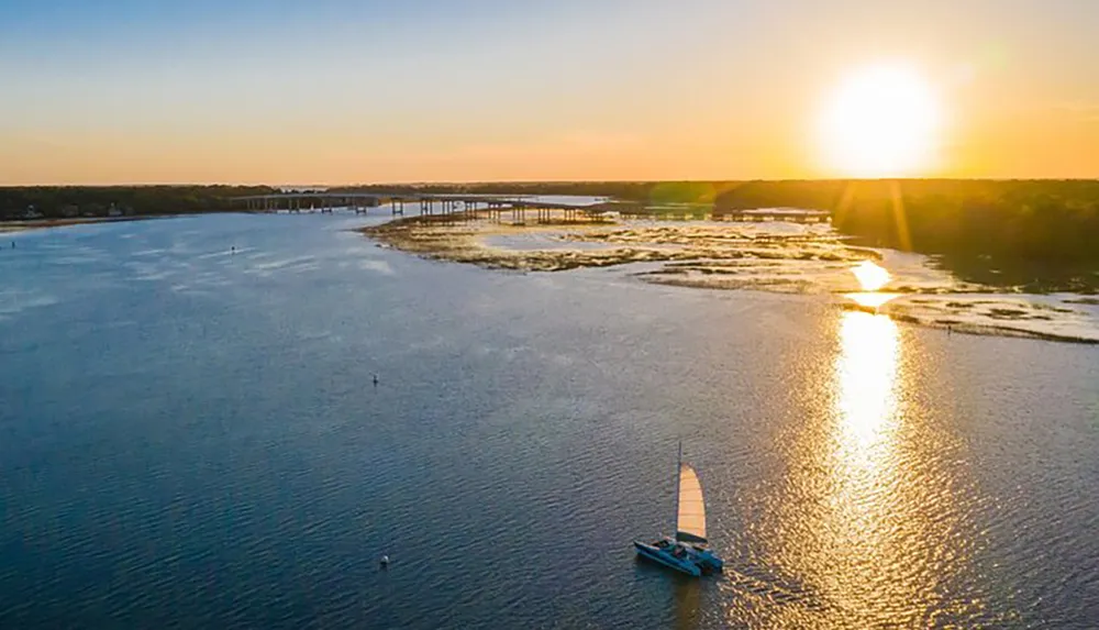 A sailboat glides on the water at sunset with a bridge in the background reflecting the warm sunlight on the rippling surface