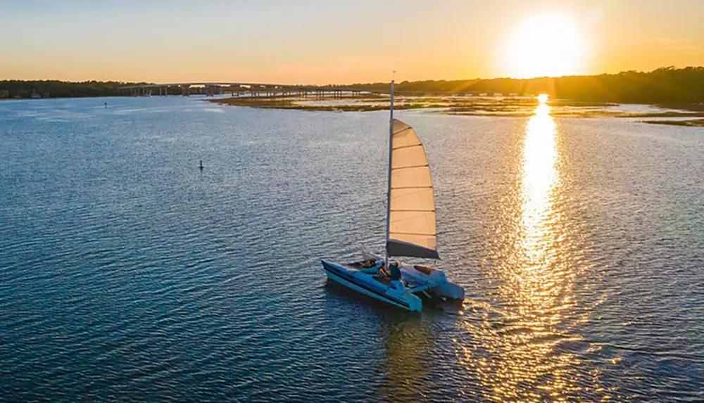 A sailboat glides on calm waters as the setting sun casts a golden path across the surface