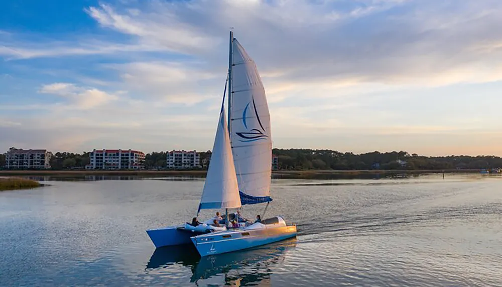 A sailboat glides across calm waters near a coastal settlement during a tranquil sunset