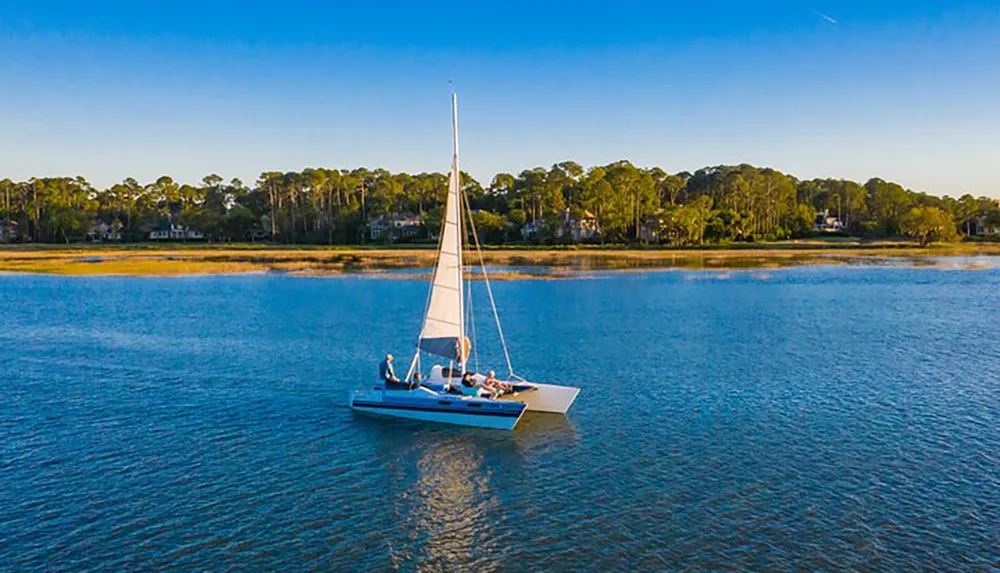 A group of people enjoys a peaceful sailboat ride on a calm blue water body near a lush tree-lined shore