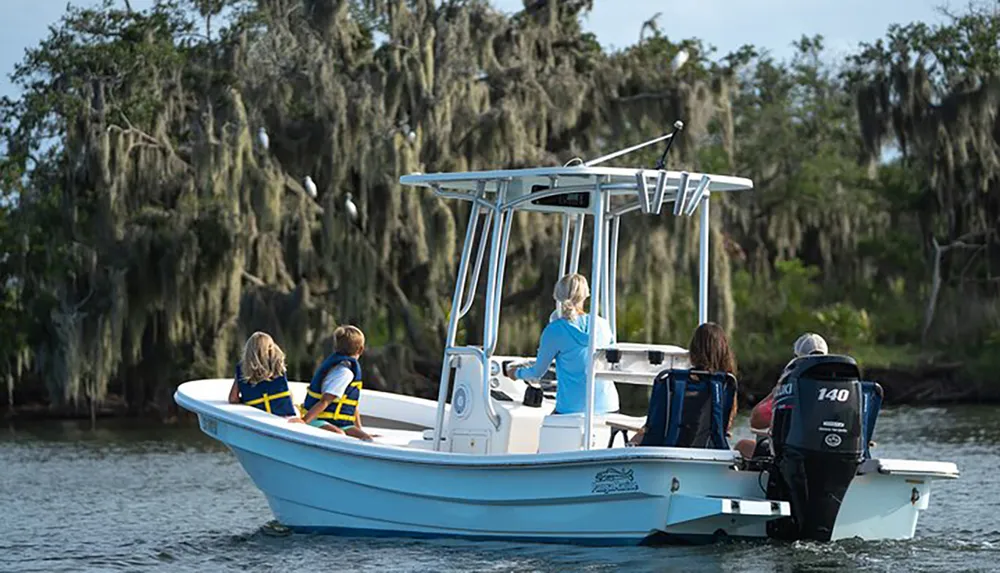 Four individuals including two children in life vests are enjoying a daytime boat ride on a tranquil river surrounded by trees draped with Spanish moss