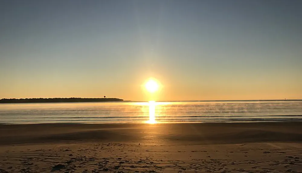 The image captures a serene sunrise over a calm ocean with the suns reflection shimmering across the water and a clear sky above a stretch of sandy beach