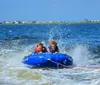 Two people are riding a blue inflatable tube on the water creating splashes as they are towed by an unseen boat