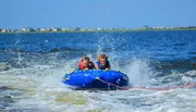 Two people are riding a blue inflatable tube on the water, creating splashes as they are towed by an unseen boat.