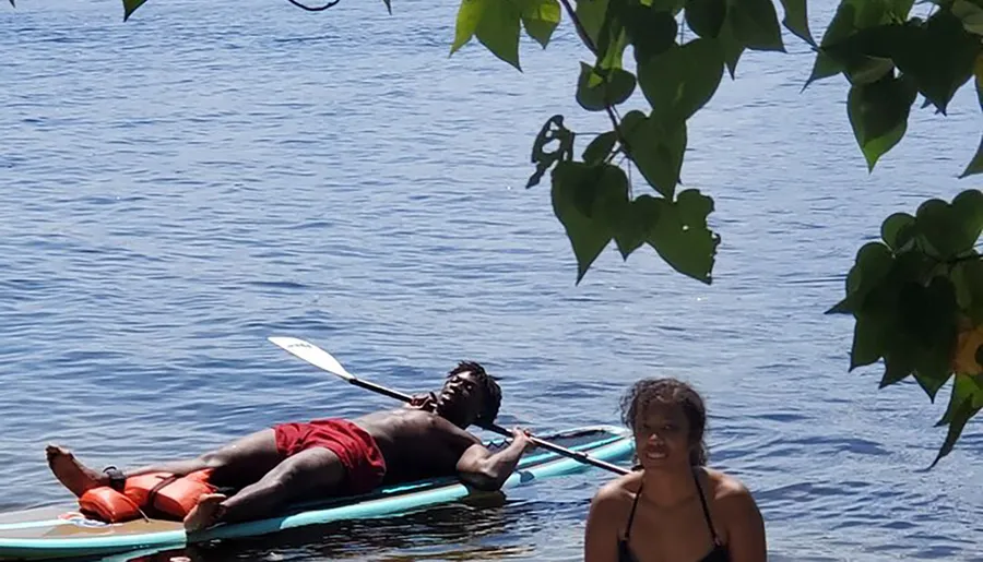 A person is lying on a paddleboard, relaxing with a paddle in hand while another individual is standing in water nearby, framed by green leaves against a backdrop of calm water.