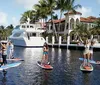 A group of people is enjoying stand-up paddleboarding in a sunny calm waterway with a luxurious mansion and a yacht in the background