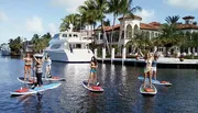 A group of people is enjoying stand-up paddleboarding in a sunny, calm waterway with a luxurious mansion and a yacht in the background.