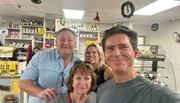 Four individuals are smiling for a selfie at a cafe or bakery, with shelves stocked with goods and Christmas decorations in the background.
