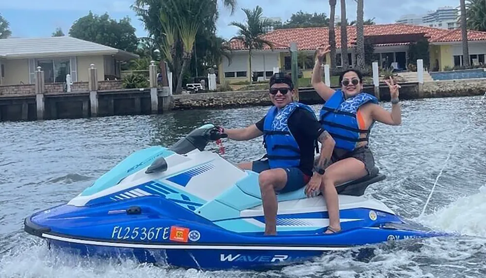 Two people are smiling and posing for a photo while riding a blue and white personal watercraft near waterfront properties with one person driving and the other raising her hand in a cheerful gesture