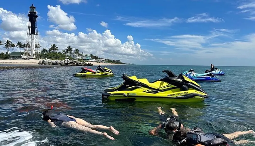 Jet skis are anchored near a rocky shoreline with people snorkeling in the foreground and an iconic lighthouse stands in the background under a partly cloudy sky