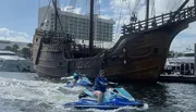 Two individuals are riding jet skis on the water near a docked replica of an old sailing ship.