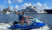 Two people are riding a jet ski with arms raised in celebration against a backdrop of yachts and water under a partly cloudy sky.