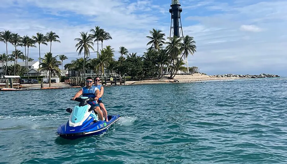 Two people are riding a jet ski near a coastline with a lighthouse and palm trees in the background