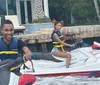 Two people wearing life vests are smiling and riding separate jet skis near a waterfront property