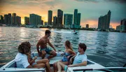 A group of friends is enjoying a boat ride against a backdrop of a vibrant sunset and the city skyline.