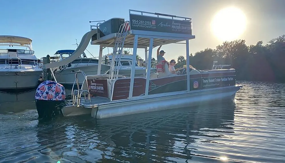 A pontoon boat with passengers is floating on calm water near other boats under a setting sun