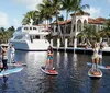 Four people are standing on paddleboards in a waterfront canal with a luxury yacht and upscale residence in the background