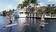 Four people are standing on paddleboards in a waterfront canal with a luxury yacht and upscale residence in the background.