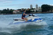 A person is riding a jet ski on the water with coastal buildings and greenery in the background.