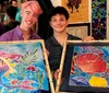 Two individuals are holding up colorful paintings of fish with smiles on their faces possibly having completed an art class or workshop