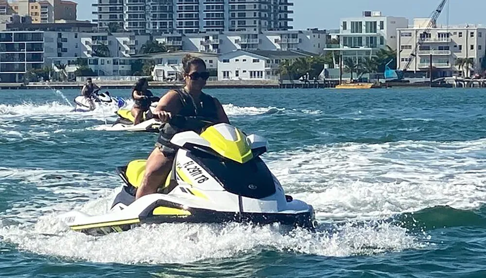 Several people are riding jet skis on a sunny day near a coastline with buildings in the background