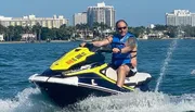 A person is riding a yellow and white jet ski on a sunny day with coastal buildings in the background.