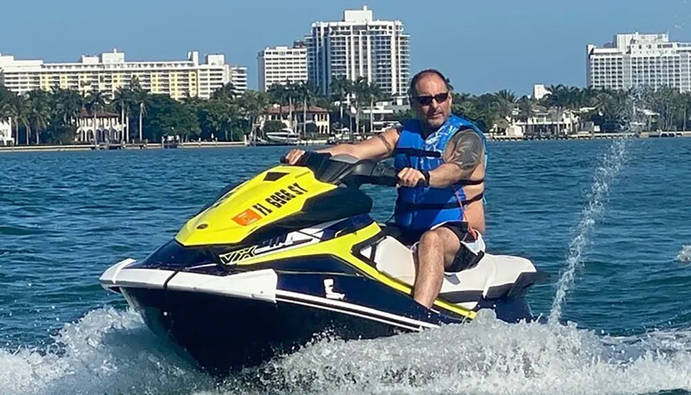 A person is riding a yellow and white jet ski on a sunny day with coastal buildings in the background