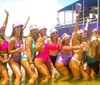 A group of people in swimwear is cheerfully posing in shallow water with drinks in their hands wearing matching visors and some of them are holding up a cutout of a persons face