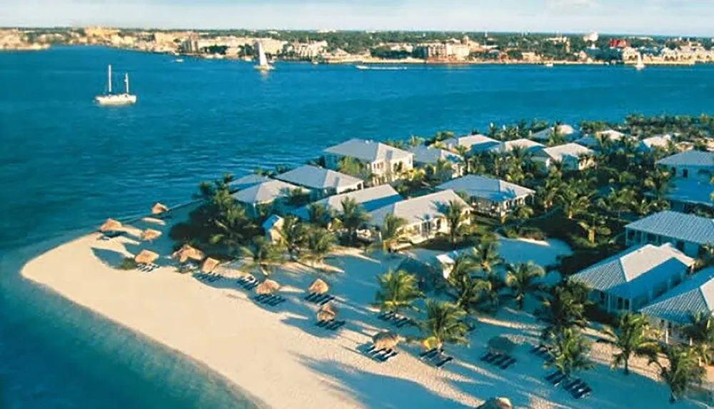 A tropical resort is nestled on a sandy peninsula with thatched umbrellas along the beach and a sailboat floating nearby in the clear blue water