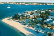 The image shows a tropical beach resort with palm trees and thatched umbrellas along a sandy shore, overlooking clear blue waters with a sailboat in the distance.